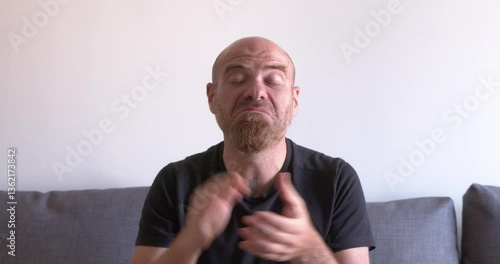 A bald young man wearing a black shirt sits on the couch and claps ironically, expressing sarcasm or mockery with a playful attitude
