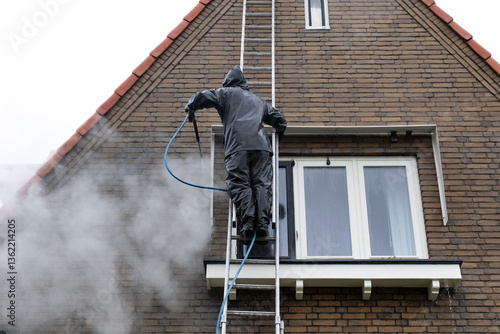 Man on ladder in waterproof suit cleans dirty brick facade of house with high pressure water jet. High pressure facade cleaning