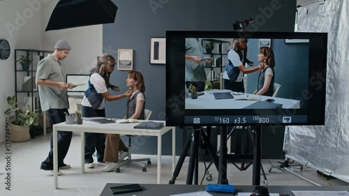 Studio monitor displaying live feed of actress at desk having her make-up adjusted, man walking by with papers, amidst professional lighting and cameras