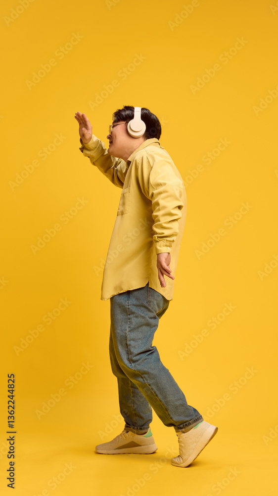 Naklejka premium Young Asian man, dressed stylish casual attire and glasses, dancing while listening music in headphones against sunny yellow background. Concept of human emotions, fashion, style, self-expression. Ad