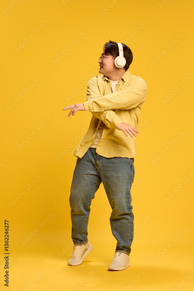 Fototapeta premium Dynamic portrait of young Asian man dressed casual, dancing while listening music, exude joy and happiness against sunny yellow background. Concept of human emotions, music, confidence, style. Ad