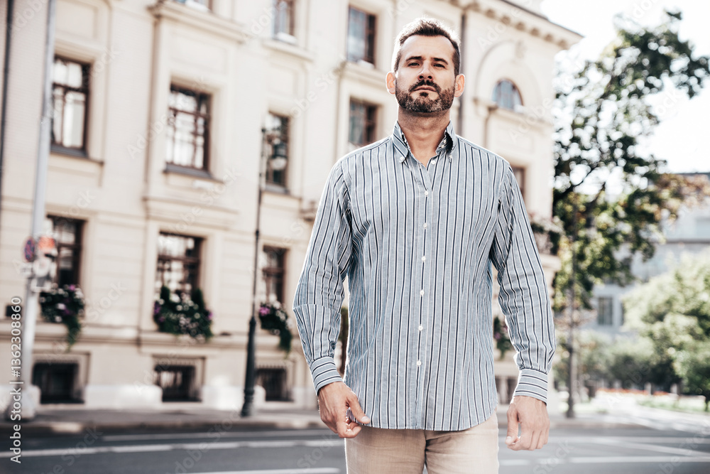 Fototapeta premium Portrait of handsome confident stylish hipster lambersexual model. Sexy modern man dressed in blue shirt and trousers. Fashion male posing on street background in Europe city at sunset