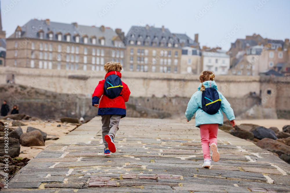 Two happy children run on a seaside stone pathway in Saint-Malo