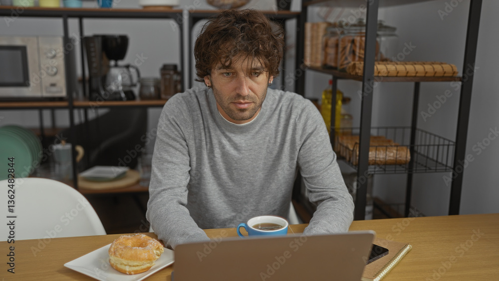 Fototapeta premium Young man working in office wearing casual sweater with coffee and donut on desk, looking focused while using laptop in organized workspace with shelves and kitchen items visible