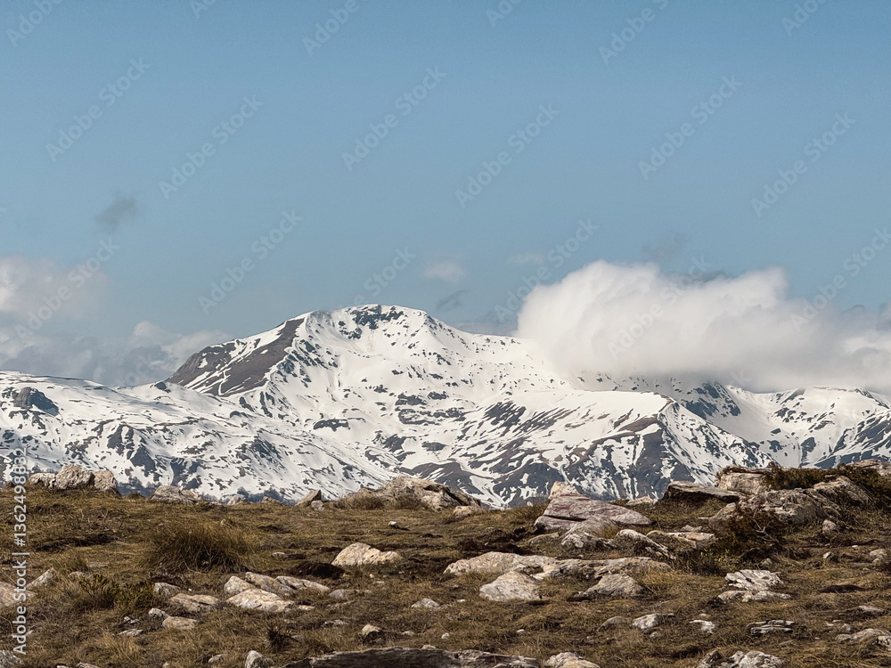 Obraz premium Hikers navigate the rugged terrain of Mavrovo National Park in Galichnik, surrounded by stunning snow-capped peaks under a clear blue sky. The atmosphere is tranquil and inviting for adventurers