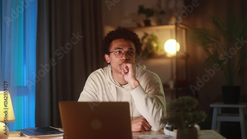 Thoughtful young man sitting at laptop at home in the evening, raising hand while thinking about ideas, options or solutions, pausing to reflect during creative or decision-making process
