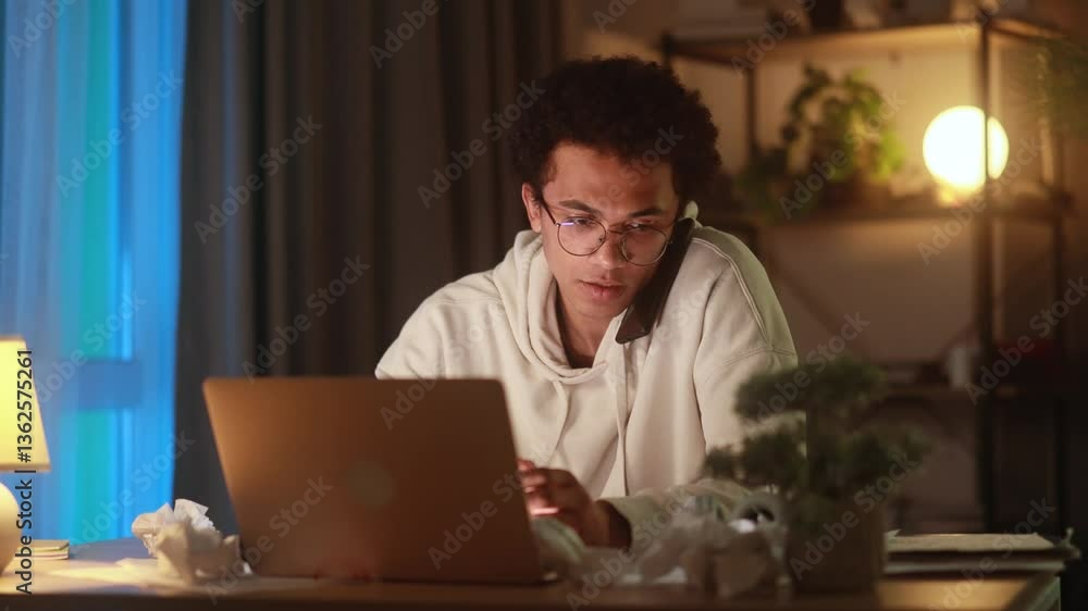 Stressed young man holding head in frustration while nervously speaking on phone and looking at laptop dealing with overwhelming situation mistake or urgent problem late at night at home Stressed young man holding head in frustration while nervously speaking on phone and looking at laptop dealing with overwhelming situation mistake or urgent problem late at night at home