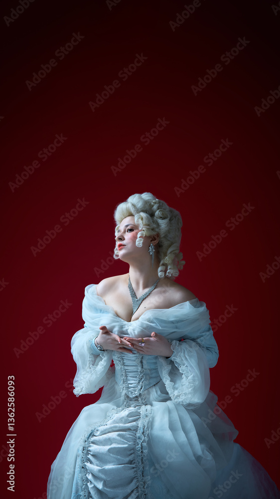 Fototapeta premium Woman in historical gown rests hands on chest, gazing upward with expressive emotion on red backdrop. Concept of historical portrait, medieval times, character.