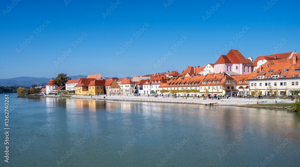 Obraz premium Picturesque view of Maribor city with the promenade and beautiful old buildings along Drava river, Slovenia.Panoramic view of the city architecture along riverbanks..