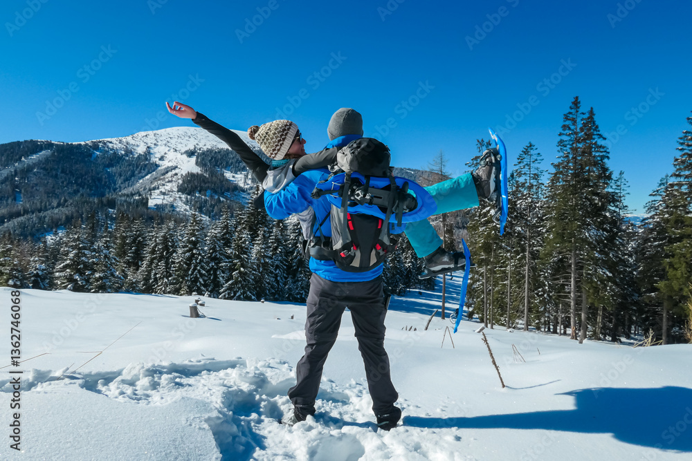 Man holding woman with snowshoes enjoying sunny winter day atop snow-covered Wölkerkogel in Styria, Austria. Panoramic view of majestic snow-capped mountain peaks in Lavanttal Alps. Snowshoe hiking