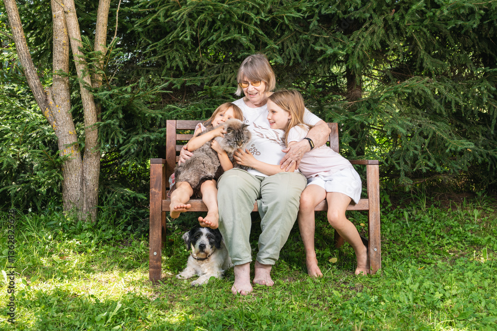 Obraz premium Grandmother sitting on a wooden bench hugging granddaughters with pet cat and dog in a summer garden