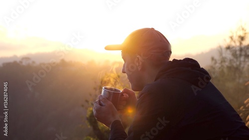 Backlit by the golden rays of the rising sun. A young man wearing a cap and casual clothes savors a hot drink. Possibly coffee or tea. While enjoying a breathtaking view of rolling hills and valleys