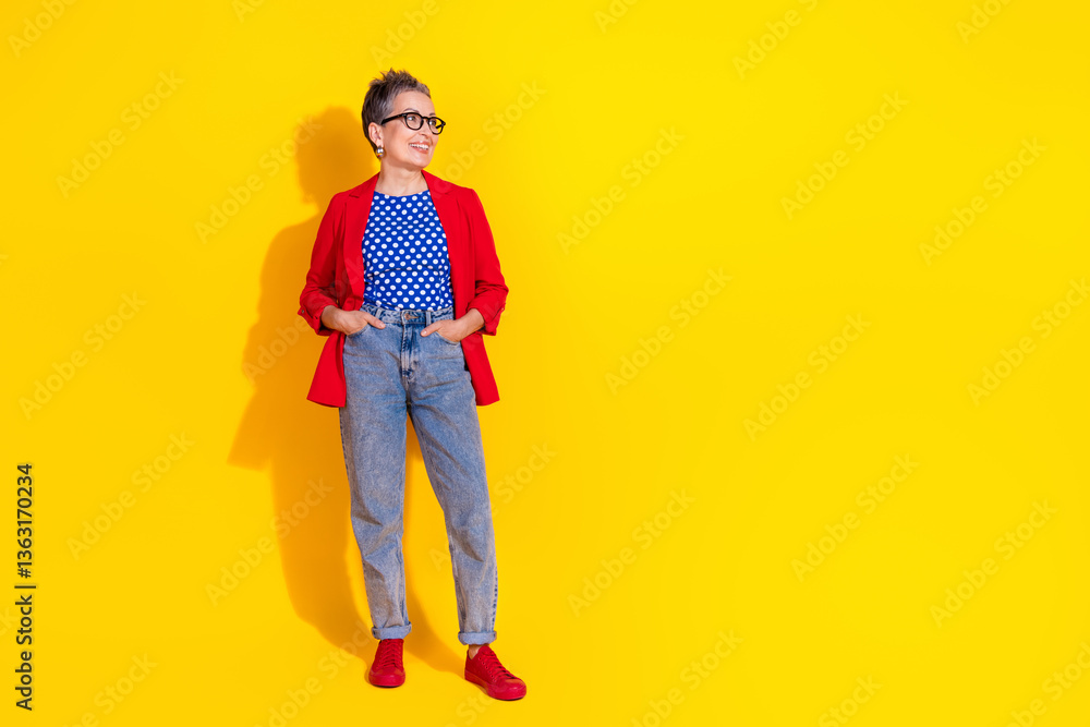 Fototapeta premium Stylish mature woman in red blazer and casual jeans posing joyfully against a vibrant yellow backdrop, expressing charm and elegance