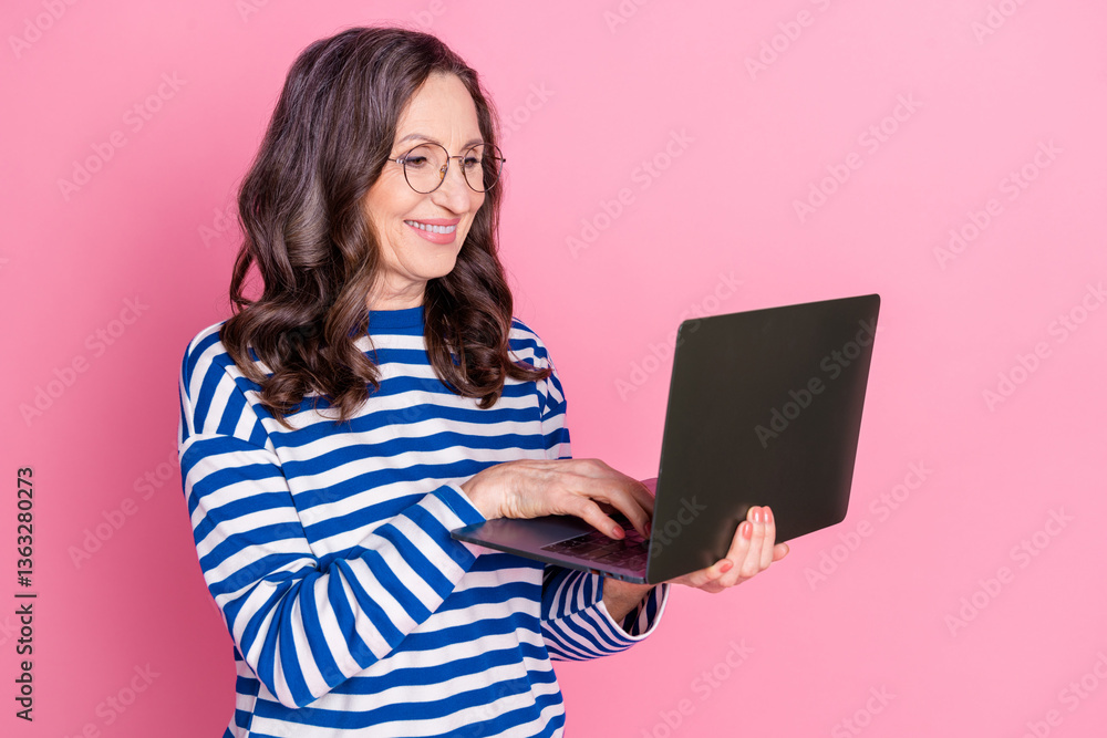 Smiling mature woman in a striped sweater using a laptop against a pink background, enjoying technology Smiling mature woman in a striped sweater using a laptop against a pink background, enjoying technology