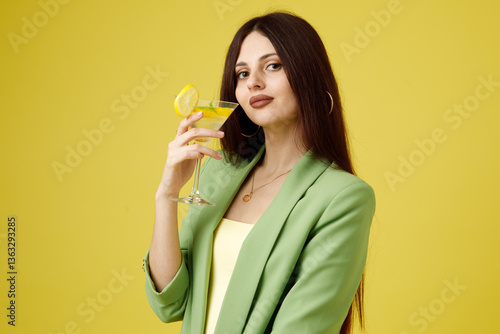 Fashion portrait of beautiful model in green suit with glass of lemonade against yellow background