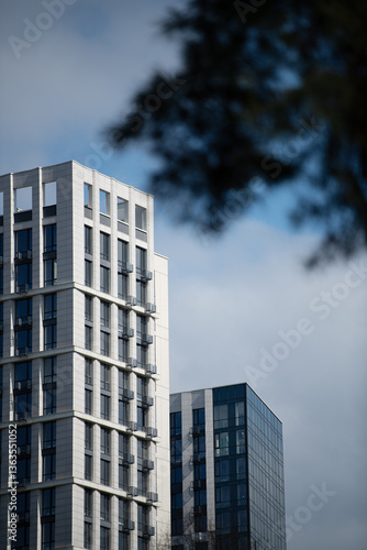 Two towering modern buildings feature striking designs, standing tall under a cloudy sky in an urban landscape. The structures display a mix of glass and concrete elements