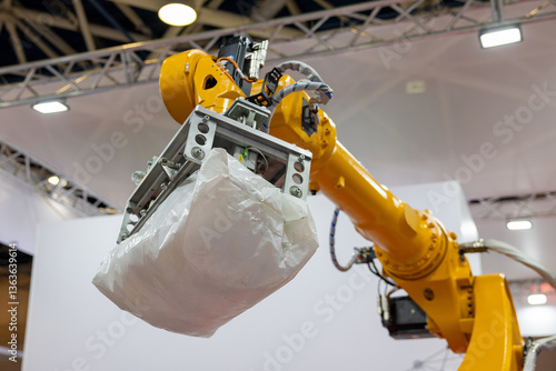An industrial robotic arm with a pneumatic gripper lifts a heavy white bag in a factory setting. The background shows a manufacturing environment. Selected focus.