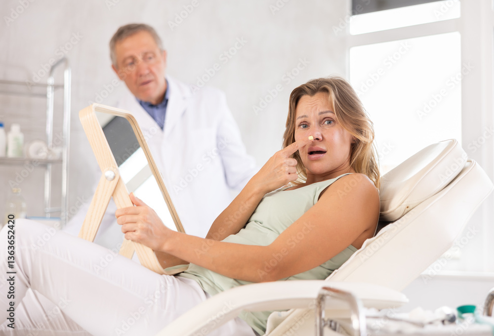 Obraz premium Upset adult woman examines her face in mirror after cosmetic procedure