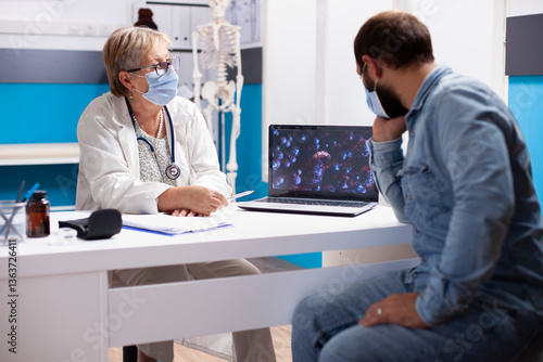 Wallpaper Mural Senior doctor seated at desk with laptop displaying virus cells and discussing with male patient in modern clinic. Old female physician and young man using digital device during medical consultation. Torontodigital.ca