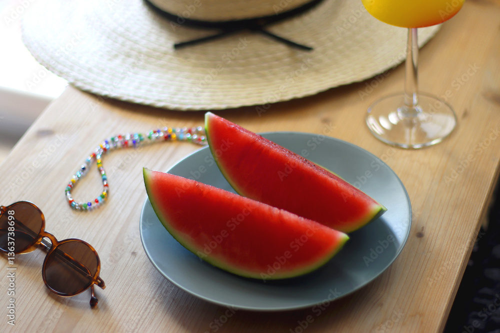 Plate with watermelon slices, glass of orange juice or cocktail, straw hat, colorful beaded necklace and sunglasses on the wooden table. Summer essentials concept. Selective focus.