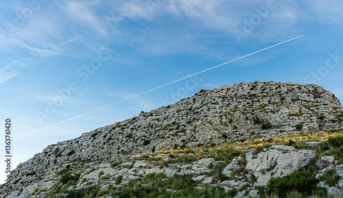 Rocky mountain landscape under a clear blue sky with scattered vegetation and a distant airplane trail