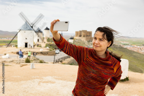 Caucasian woman standing at Spanish windmills of Consuerga and taking selfies with her smartphone.