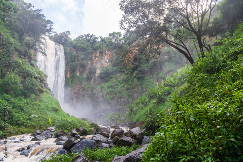 Thompson Falls in Kenya is a magnificent waterfall plunging down a steep cliff into a pool below, surrounded by lush green vegetation and rocky outcrops. Mist rises from the base, creating a serene