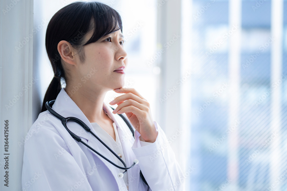 A female doctor leans against a window, hand on chin, gazing into the distance in deep thought. She wears a white coat and stethoscope, standing in a brightly lit medical facility.