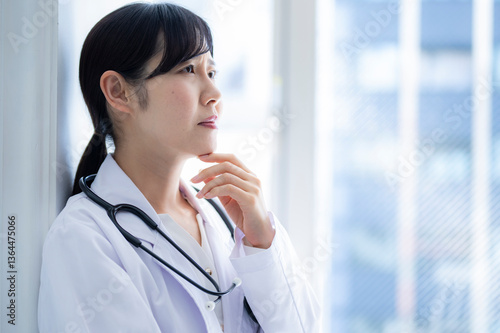 A female doctor leans against a window, hand on chin, gazing into the distance in deep thought. She wears a white coat and stethoscope, standing in a brightly lit medical facility.