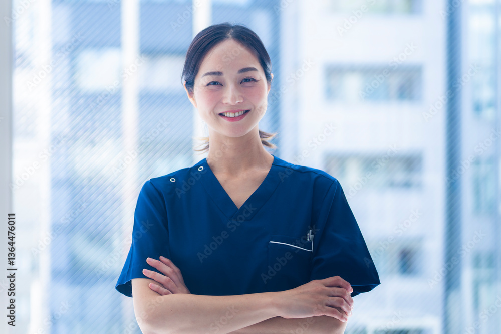 A young female nurse in blue scrubs stands confidently with folded arms inside a modern hospital building. She is smiling warmly, suggesting professionalism and compassion.