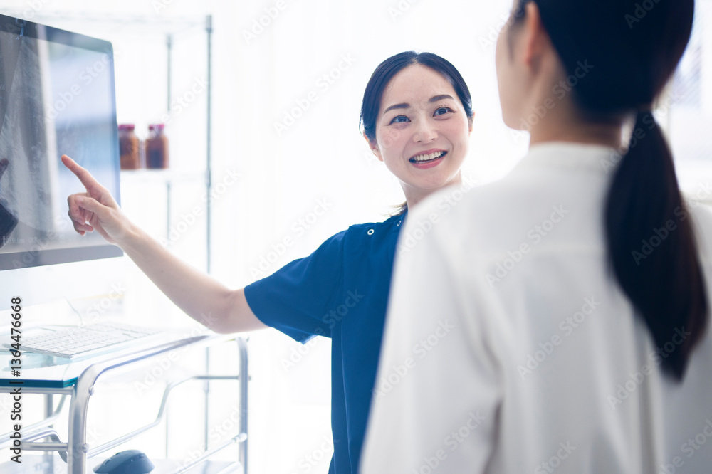 A female doctor in blue scrubs is smiling and pointing at a medical monitor while explaining results to a patient in a clinical setting.