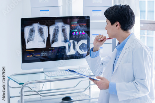 A young male doctor in a white lab coat is deeply focused while reviewing a patient's chest X-ray and analytical graphs displayed on a computer monitor. He is holding a blue clipboard and a pen, worki