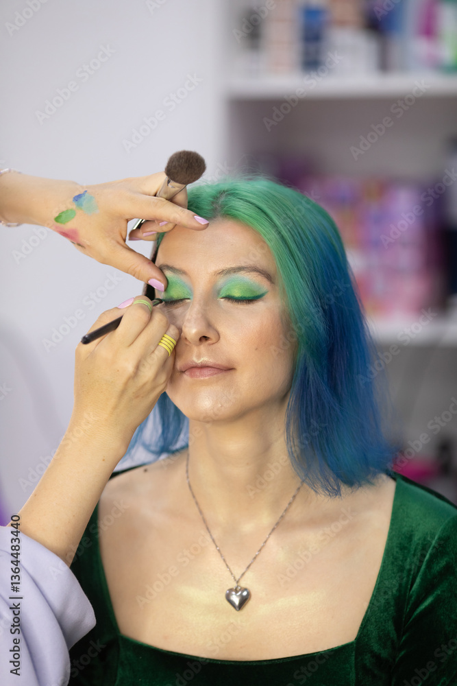 Fototapeta premium woman with green hair is getting her eyes done by a stylist. The stylist is using a brush to apply makeup to the woman's eyes. The woman is relaxed and comfortable with the process