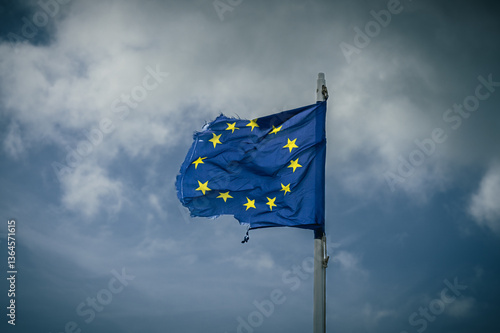 A torn EU flag blows in the wind with a few clouds in the background