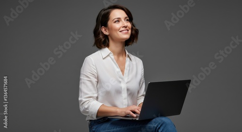 Woman in white shirt using laptop and smiling while looking up against gray background in studio shot