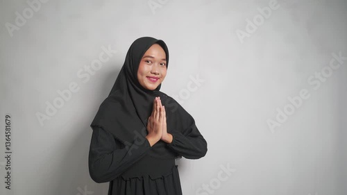 A smiling asian woman wearing a black hijab with her hands together in a respectful gesture.