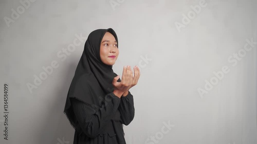 A young asian woman in black hijab gazes thoughtfully, set against a plain grey backdrop.