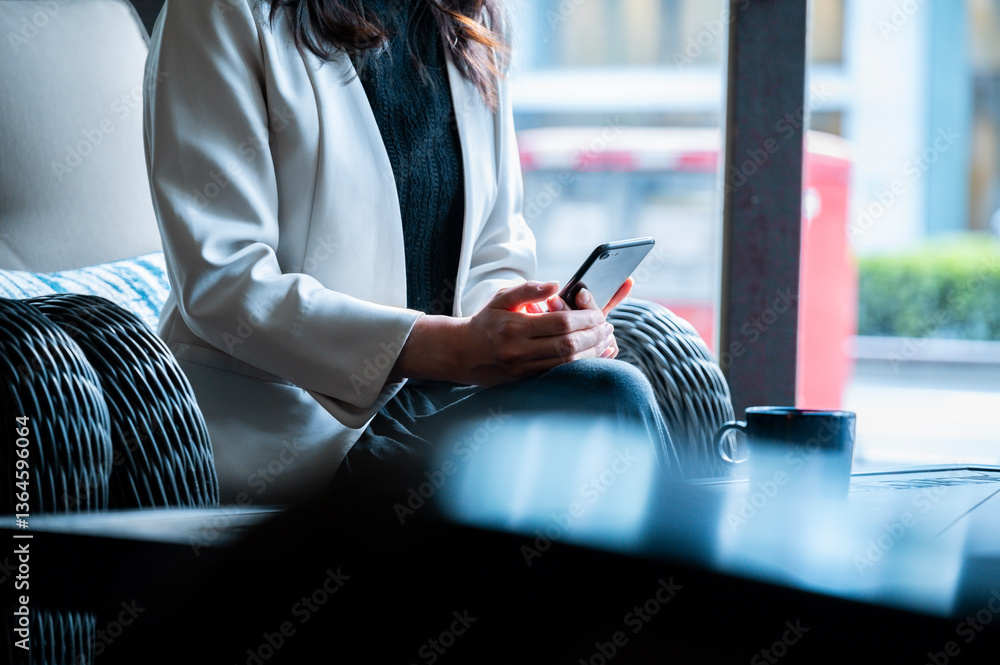 A woman in a light blazer sits comfortably in a stylish café, holding a smartphone with both hands. The setting includes a woven armchair, a patterned cushion, a coffee mug, and natural daylight filte