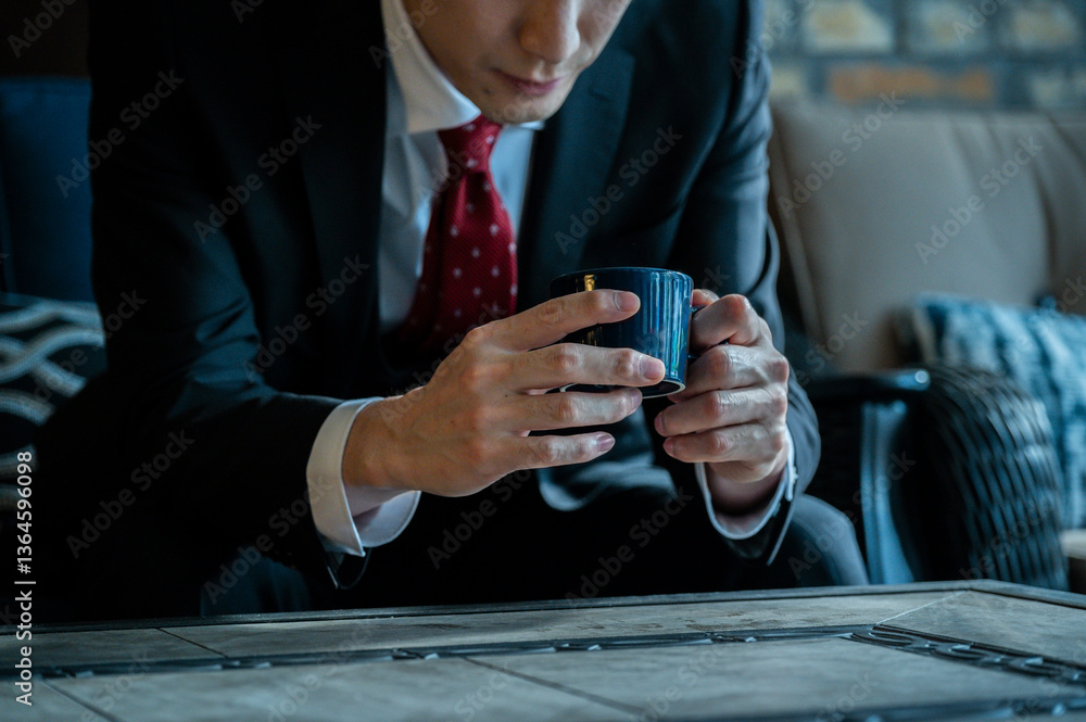 A man in a black suit and red tie sits on a wicker chair near a large window holding a coffee cup, appearing relaxed and thoughtful.