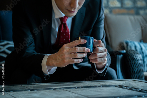Obraz na płótnie A man in a black suit and red tie sits on a wicker chair near a large window holding a coffee cup, appearing relaxed and thoughtful. w salonie