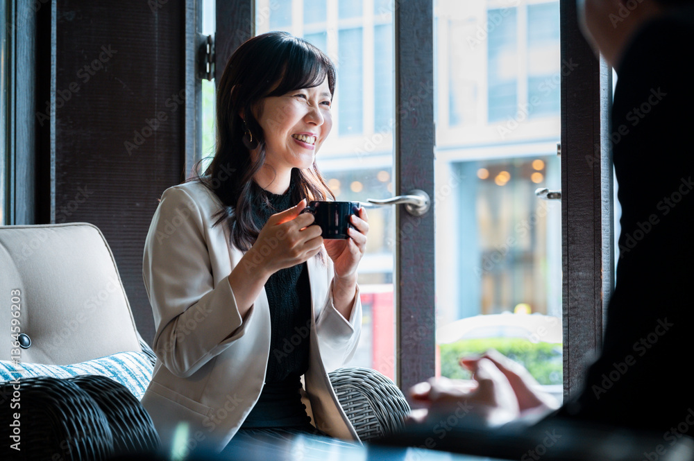 cheerful woman in a cream blazer enjoys a cup of coffee while seated by a window in a stylish café. Natural daylight highlights her relaxed mood as she looks toward someone outside the frame.