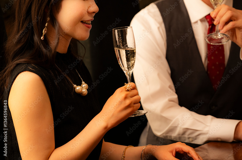 A man in a white shirt and vest and a woman in a black dress toast with champagne glasses at a high-end bar. The couple is smiling and enjoying a relaxed evening in a dimly lit, elegant setting.