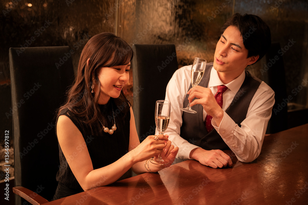 A man and woman dressed formally are smiling at each other while toasting with champagne glasses. They are seated across from each other at a wooden bar with warm ambient lighting.