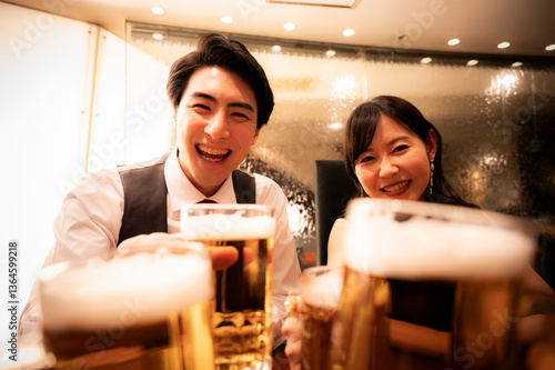 Obraz na płótnie A smiling couple raises their glasses of beer with friends in a bright, warmly lit bar. The mood is festive and welcoming, full of laughter and joy. w salonie