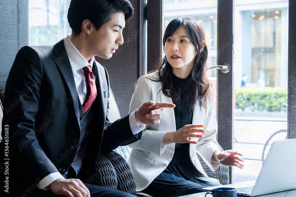 A businessman and a businesswoman in formal attire have a serious conversation while seated by the window at a modern café. Natural light highlights their focused expressions as they engage in a profe