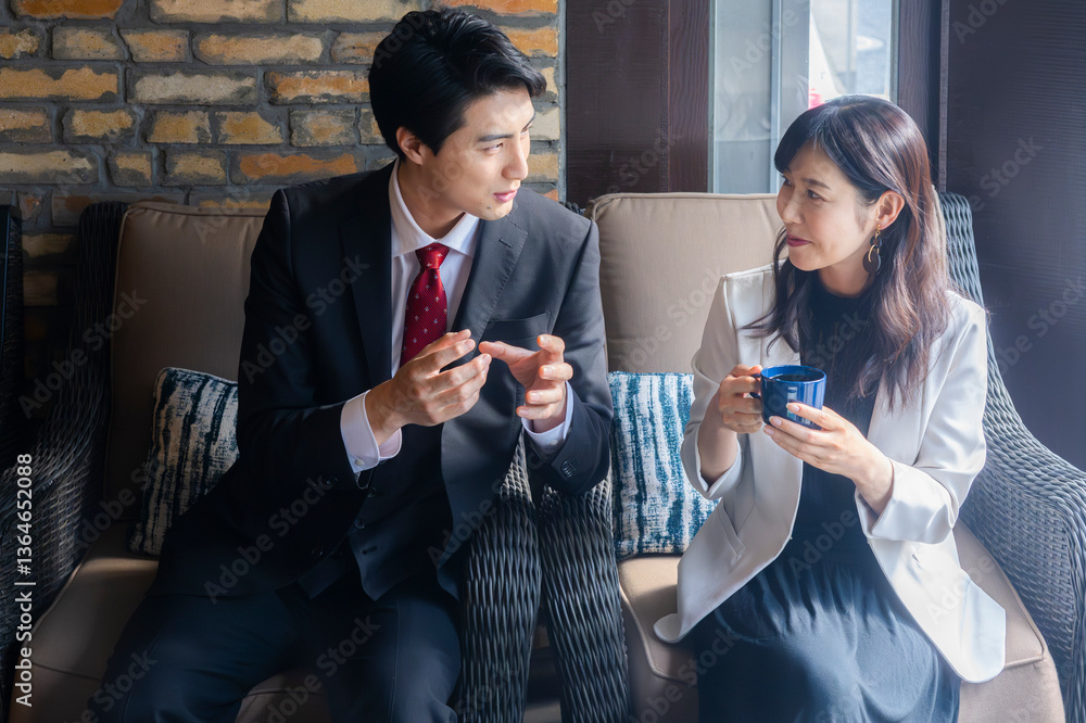 A businessman in a dark suit and red tie gestures while speaking with a female colleague in a light blazer during a strategic meeting in a cozy café with a brick wall background.