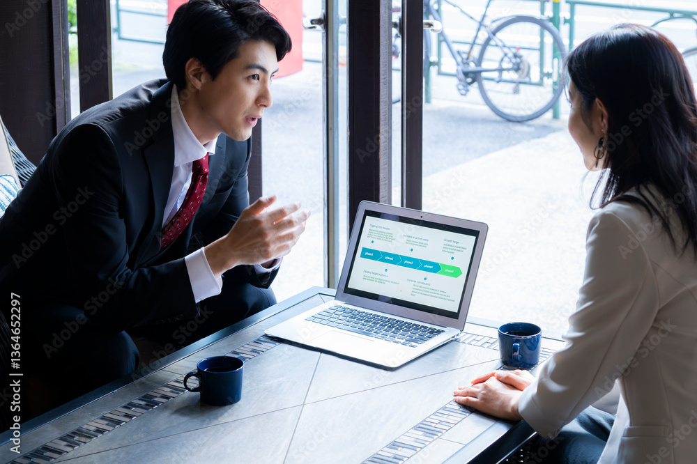 A businessman in formal attire is pointing to a laptop screen and explaining data to a female colleague across the table. They are seated by a bright window in a stylish café setting with a coffee mug