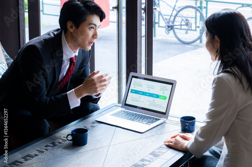 Obraz na płótnie A businessman in formal attire is pointing to a laptop screen and explaining data to a female colleague across the table. They are seated by a bright window in a stylish café setting with a coffee mug w salonie