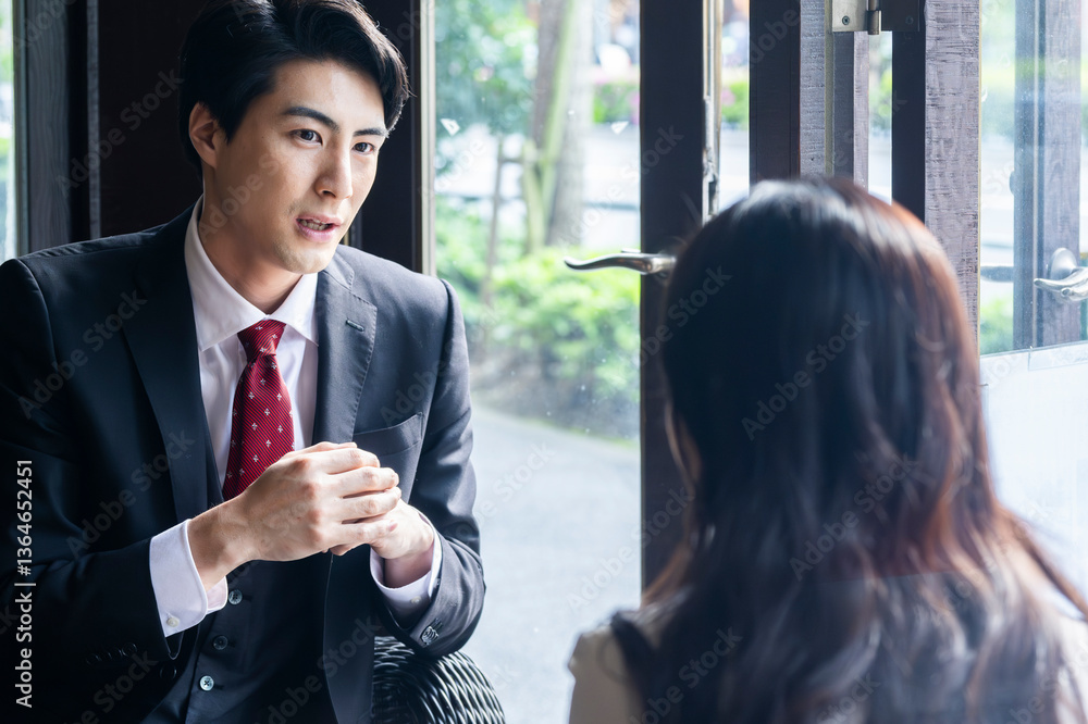 A businessman in formal attire is pointing to a laptop screen and explaining data to a female colleague across the table. They are seated by a bright window in a stylish café setting with a coffee mug