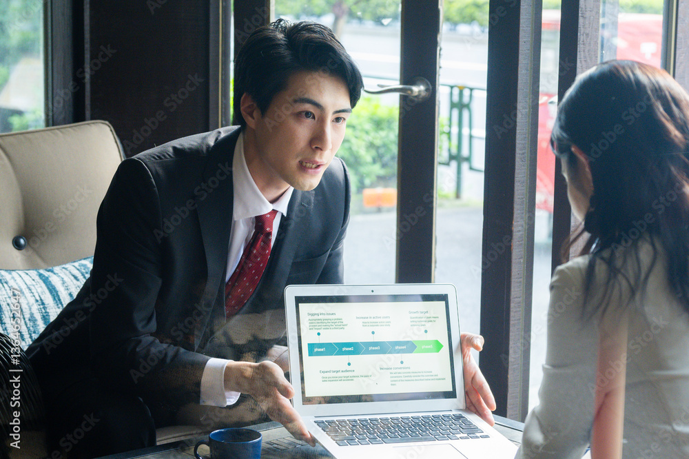 A businessman in formal attire is pointing to a laptop screen and explaining data to a female colleague across the table. They are seated by a bright window in a stylish café setting with a coffee mug